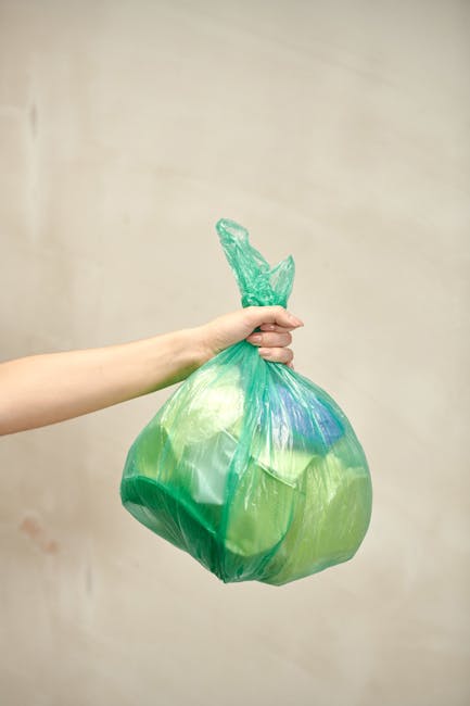 A person's hand is visible holding a green plastic rubbish bag tightly by its tied top. The bag appears to be full, with some bulging areas suggesting contents inside. The plastic has a shiny, smooth texture and the bag includes various shades of green, with slight reflections indicating a light source nearby. The background is a plain, light-colored wall, with no additional objects or environmental details visible. The scene emphasizes the act of waste collection or disposal, aligning with services such as independent rubbish removal or on-site waste handling provided by Rubbish Collection Finchley. The focus is on the bag's material, color, and the action of holding it, which highlights the process of rubbish disposal in a straightforward, professional manner suitable for accessibility and SEO considerations relevant to private waste collection services.