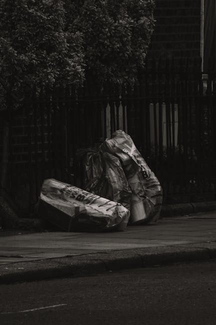 Three large black plastic garbage bags filled with waste are placed on a paved sidewalk next to a dark metal fence and an area with dense foliage. The bags appear to contain household rubbish or similar waste materials and are slightly crumpled, with some showing text printed on the surfaces. They are positioned close to the curb, on a street that appears to be in an urban or residential setting, with the background comprising the fence and overhanging trees. The scene is illuminated by natural ambient light, creating subtle highlights and shadows on the bags and surrounding environment. This image visually relates to private or independent rubbish collection services, such as those offered by rubbish removal companies like Rubbish Collection Finchley, emphasizing the need for professional waste handling and disposal solutions outside local authority services.