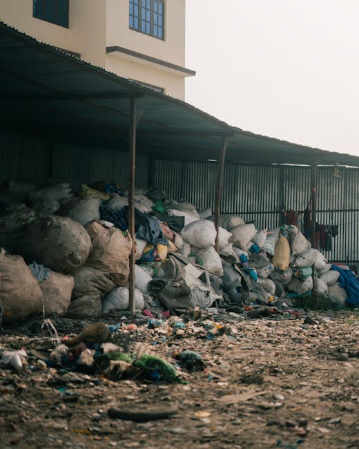 A collection of overflowing rubbish and waste bags piled around a large, grey, metal communal waste bin situated on a paved urban street. The waste includes black and blue bin bags, flattened cardboard boxes, crumpled paper, plastic bottles, and loose packaging materials, some spilling onto the surrounding pavement and curb area. To the left of the main waste container, there are additional cardboard boxes and miscellaneous debris. Behind the waste area, a grey vehicle with a yellow and black license plate is parked parallel to a metal railing that separates the waste from the sidewalk. The background features a modern building under construction, wrapped in blue protective scaffolding, with signage for local businesses visible below. The scene appears to depict an example of poorly managed or uncollected rubbish, highlighting the importance of regular waste disposal or alternative collection services like those provided by Rubbish Collection Finchley, especially for private or on-site rubbish removal in urban environments.