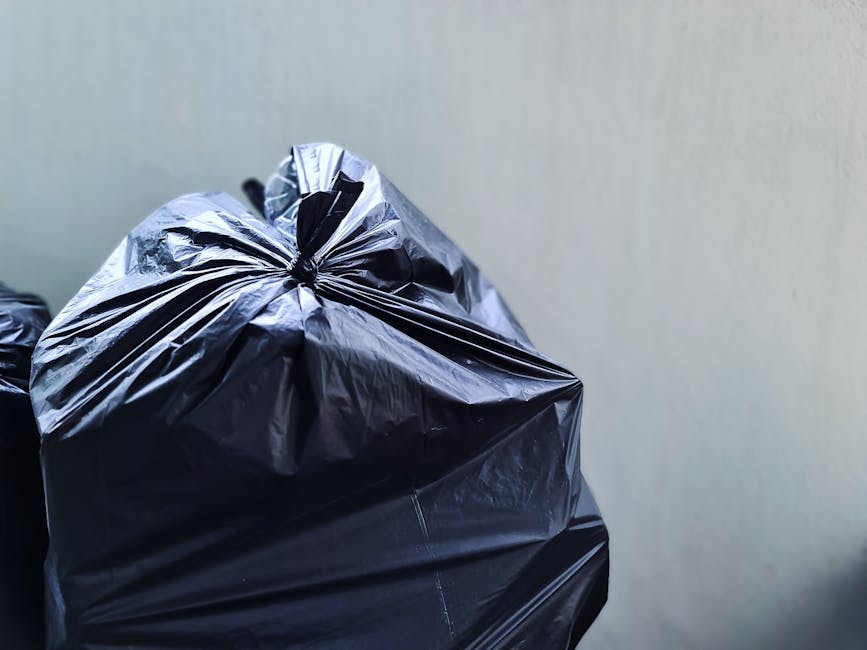 Close-up of tightly knotted black plastic rubbish bags, made from glossy, flexible material, resting against a plain, light-colored wall. The bags appear filled and bulging, with some creases and folds visible on their surface, indicating they are full of household waste. They are positioned in the lower left corner of the image, with one bag slightly overlapping another. The environment suggests an indoor or outdoor setting such as a garage, driveway, or storage area used for waste collection or disposal. The lighting is neutral and even, highlighting the texture and sheen of the plastic material. This scene relates to rubbish removal services offered by Rubbish Collection Finchley, with emphasis on the process of preparing waste for collection. The focus on the bags' condition and placement supports the context of private waste handling and on-site clearance for homeowners or property managers in the Finchley area.