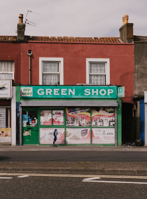 The image shows the front of a small retail shop with a bright green sign reading 'GREEN SHOP' and the smaller text 'GKV NEWS' on the left side. The shop is located on a street with a dark pavement, featuring a large window display covered with various advertisements and posters. The storefront has a green frame and a matching green door on the left, which appears to be a secondary entrance. Behind the shop, there is a two-story building with a faded pink exterior, two windows with white frames and curtains, and a roof with red tiles. Two brick chimneys are visible on either side of the roof, with weathered television antennas attached to the chimney on the left. The sky is overcast, casting diffuse light over the scene. The environment suggests a typical urban or semi-urban area, with the shop possibly involved in retail or community services, and occasional waste or rubbish collection happening nearby, as part of routine maintenance or private clearance services such as those provided by Rubbish Collection Finchley.
