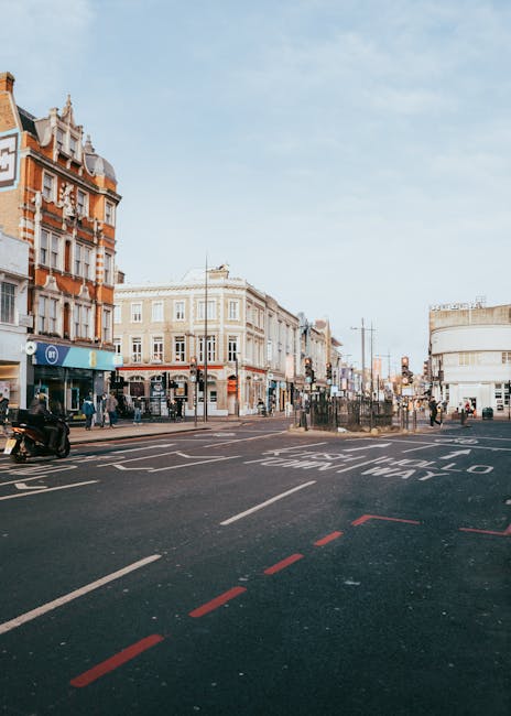 A wide view of North Finchley High Road showing a busy urban street scene during daytime, with a mixture of historic and modern buildings lining the pavement. The foreground features a section of a dark asphalt road with white lane markings and red dashed lines, indicating parking restrictions or loading zones. To the left, there is a black scooter parked, and pedestrians are visible walking along the sidewalk, some near small shops and retail outlets. In the background, there are more pedestrians crossing or standing near shop fronts, with streetlights and signage visible further along the street. The sky is clear with a few clouds, and the overall scene depicts a typical shopping district with active foot traffic, suitable for services related to waste or rubbish collection supplied by local waste management providers such as Rubbish Collection Finchley, operating in the area.