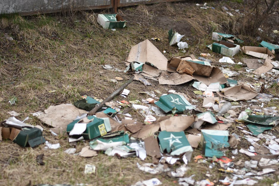An outdoor scene showing an uneven grassy area next to a paved surface, with scattered debris including torn and crumpled cardboard boxes, some with printed branding or logos, and pieces of paper and plastic waste. The cardboard items are in various shades of brown and green, some partially flattened or broken. Flat, weathered cardboard sheets are mixed with smaller trash fragments, all spread across the ground. A metal pole is also visible among the debris, lying diagonally on the ground, partially covered by the scattered rubbish. The background includes a section of a wooden fence and overgrown vegetation, with dry branches and sparse grass evident throughout the scene. The lighting appears natural, suggesting daylight, and the overall setting reflects an area affected by waste dumping, characteristic of a site requiring professional rubbish collection or on-site clearance, as practiced by Rubbish Collection Finchley.