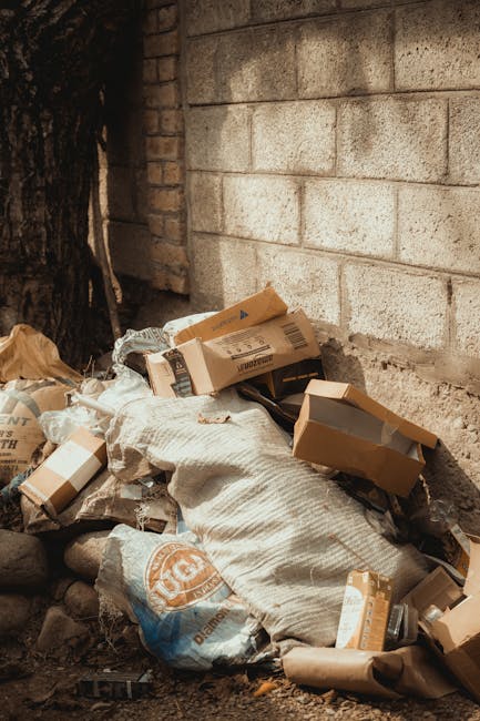An outdoor scene showing a pile of mixed rubbish including flattened cardboard boxes, plastic bags, and paper packaging, some of which are torn or crumpled. The waste is situated directly against a weathered brick wall with visible mortar lines, and partially shaded by a nearby tree trunk on the left side of the image. The cardboard boxes vary in size and are mostly brown, with some featuring printed labels or logos, while the plastic bags appear semi-transparent or opaque in shades of beige and white. The ground beneath the rubbish consists of dirt and small stones, with a few scattered leaves. The scene suggests an on-site private waste collection or rubbish removal, typical of residential or commercial refuse clearance, with a focus on easy access for collection by waste removal services like Rubbish Collection Finchley, which offers alternative rubbish disposal options to standard curbside collection.