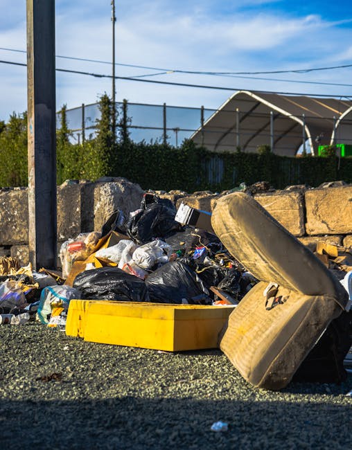 A pile of mixed waste materials is gathered at the side of a paved area, consisting of black plastic rubbish bags, a yellow plastic container, and a large, worn car tyre leaning against the bags. The waste includes various items such as paper, plastic packaging, and other debris, all disorderly arranged and spilling onto the ground. Behind the pile, there is a low stone wall made of irregularly shaped bricks or concrete blocks, with some greenery like bushes and small trees visible beyond it. In the background, a blue sky with scattered clouds is seen above overhead power lines, and a partially visible structure with a curved roof or canopy suggests an outdoor setting possibly associated with private waste collection or an on-site clearance operation. The scene appears to depict an area awaiting rubbish collection, perhaps arranged by Rubbish Collection Finchley for alternative waste handling or private disposal services.