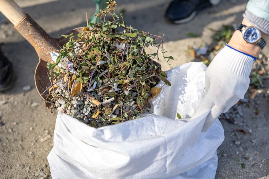 A close-up image of a person's gloved hand holding a white, woven polypropylene bag filled with garden and yard waste, including soil, small rocks, dry leaves, and plant debris. The person's wrist is visible, featuring a silver wristwatch and a light-colored, long-sleeved sweater. The bag is partially open, revealing the mixture of organic and inert materials inside. A rusted shovel with a wooden handle is positioned behind the bag, with its blade partly buried in the ground, which appears to be a gravel or dirt surface. The background shows a paved area and some indistinct foliage. The scene suggests an outdoor setting where waste collection or garden clearance is taking place, aligning with the services offered by Rubbish Collection Finchley for efficient, private rubbish removal or on-site clearance solutions. The lighting is natural, indicating daytime, with shadows cast by the objects, emphasizing the textures and materials involved in waste disposal activities.