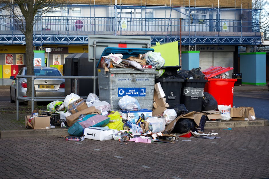 A pile of mixed waste materials is gathered at the side of a paved area, consisting of black plastic rubbish bags, a yellow plastic container, and a large, worn car tyre leaning against the bags. The waste includes various items such as paper, plastic packaging, and other debris, all disorderly arranged and spilling onto the ground. Behind the pile, there is a low stone wall made of irregularly shaped bricks or concrete blocks, with some greenery like bushes and small trees visible beyond it. In the background, a blue sky with scattered clouds is seen above overhead power lines, and a partially visible structure with a curved roof or canopy suggests an outdoor setting possibly associated with private waste collection or an on-site clearance operation. The scene appears to depict an area awaiting rubbish collection, perhaps arranged by Rubbish Collection Finchley for alternative waste handling or private disposal services.