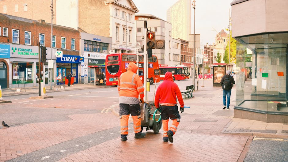 Two waste collection workers dressed in high-visibility orange and red reflective uniforms are pushing a wheeled bin along a busy city street. One worker is holding the handle of the bin, while the other guides it from the side. The street features a red double-decker bus stopped at a traffic light, with its doors open. Surrounding storefronts include a building society branch, a store named Coral, and a college, with signs visible on the shop fronts. In the background, pedestrians are walking, some nearby and others further down the street, while a traffic light displays a red signal. To the right, a glass-fronted building or kiosk is partially visible, with a few people passing by. The scene is illuminated by natural daylight, suggesting an early morning or late afternoon setting, within an urban area that may be managed by independent waste removal services such as Rubbish Collection Finchley, reflecting a typical rubbish collection or on-site clearance operation in a city environment.
