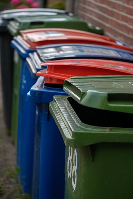 Two waste collection workers dressed in high-visibility orange and red reflective uniforms are pushing a wheeled bin along a busy city street. One worker is holding the handle of the bin, while the other guides it from the side. The street features a red double-decker bus stopped at a traffic light, with its doors open. Surrounding storefronts include a building society branch, a store named Coral, and a college, with signs visible on the shop fronts. In the background, pedestrians are walking, some nearby and others further down the street, while a traffic light displays a red signal. To the right, a glass-fronted building or kiosk is partially visible, with a few people passing by. The scene is illuminated by natural daylight, suggesting an early morning or late afternoon setting, within an urban area that may be managed by independent waste removal services such as Rubbish Collection Finchley, reflecting a typical rubbish collection or on-site clearance operation in a city environment.